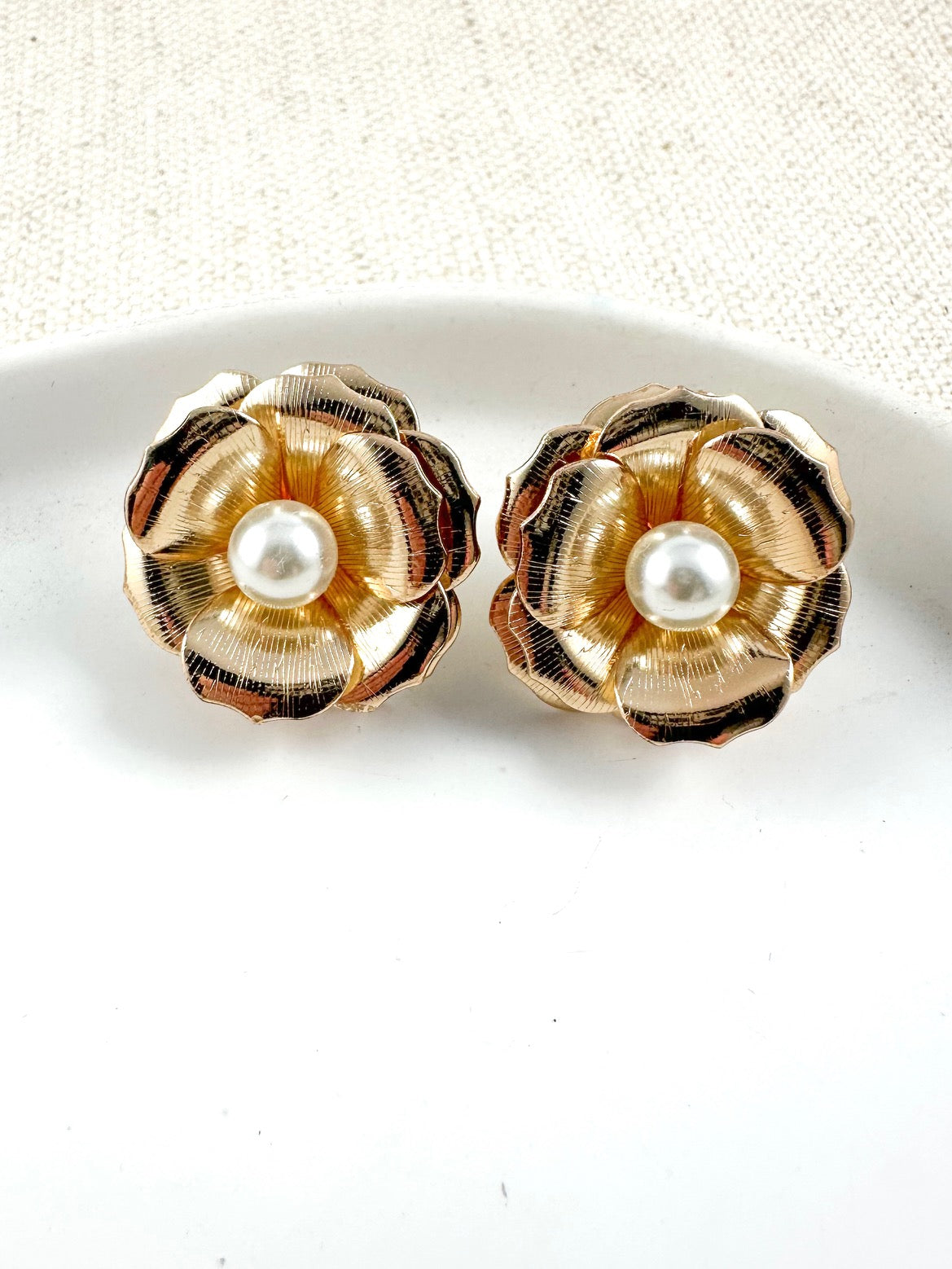 Gold floral stud earrings with textured petals and a pearl center on a light background.
