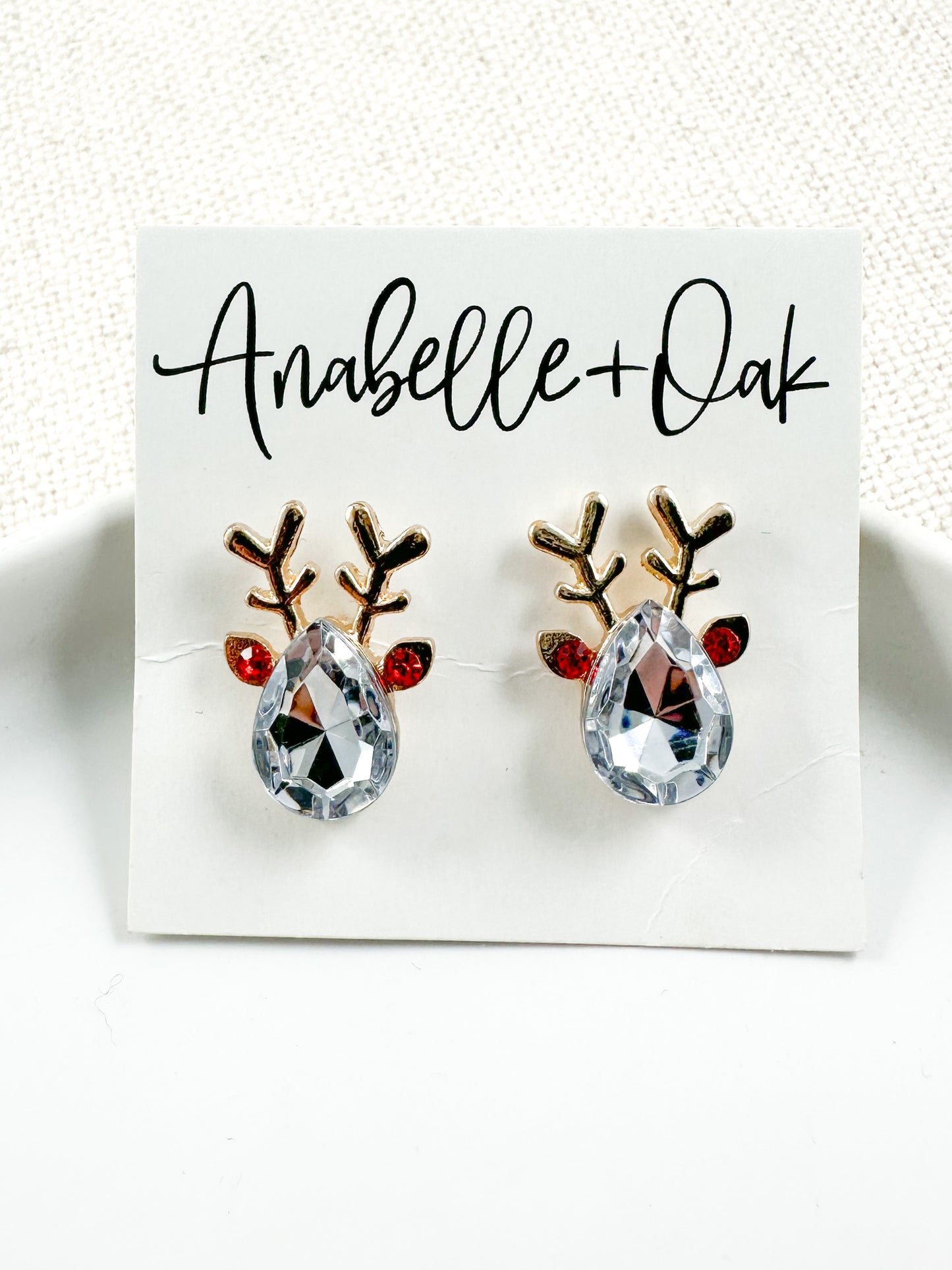 Gold reindeer stud earrings with clear teardrop crystals and red crystal accents on an Anabelle+Oak earring card, styled on a white dish with a neutral background.