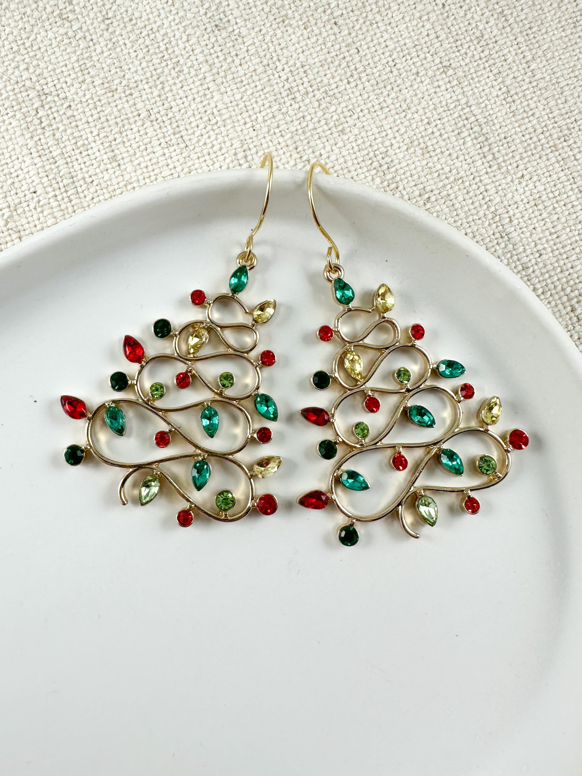 A pair of gold-colored earrings with red and green crystal embellishments, displayed on a white plate against a textured background.