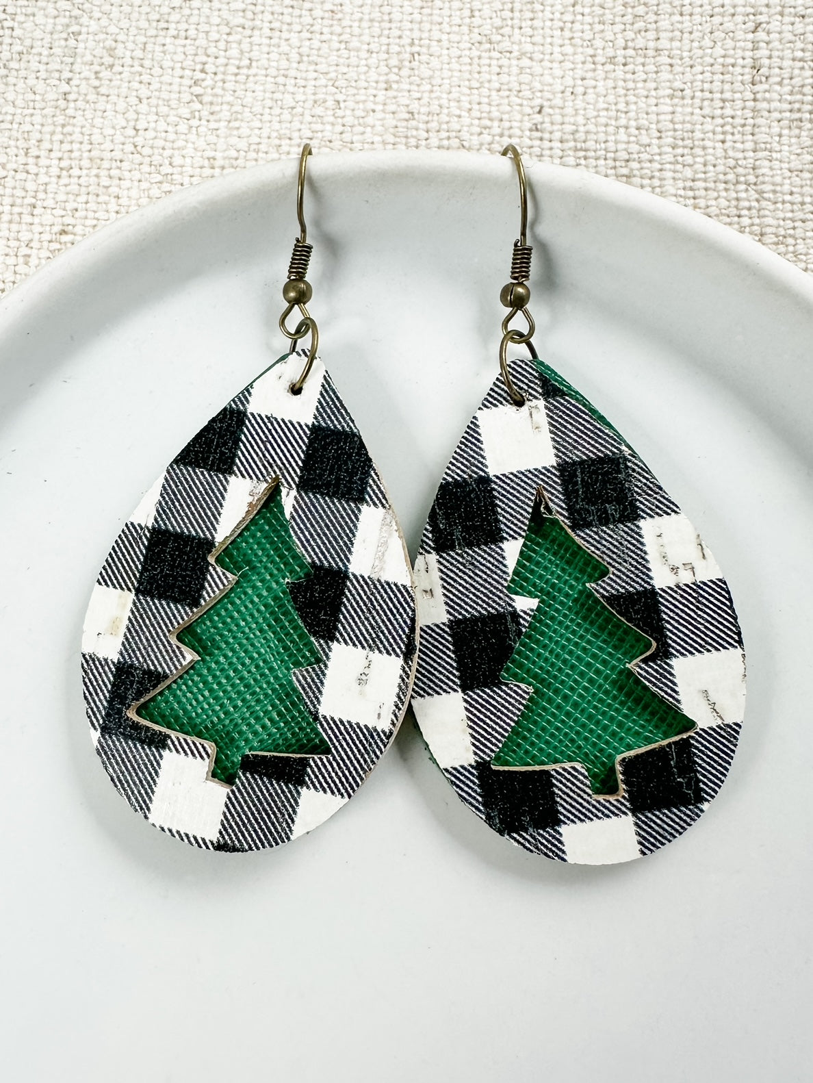 A pair of earrings featuring a black and white checkered pattern with a green Christmas tree cutout, displayed on a white plate.