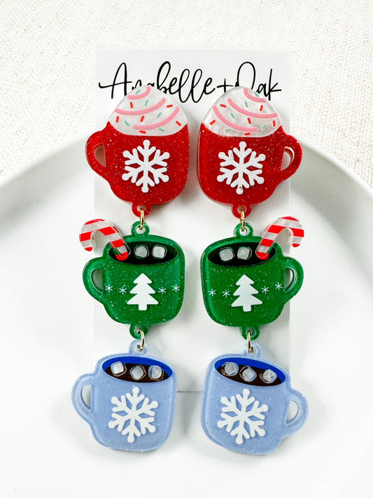 Festive acrylic Christmas cocoa mug earrings featuring red, green, and blue glitter mugs with snowflake and tree designs, candy cane accents, and whipped cream details — lightweight 3" holiday statement earrings from Anabelle+Oak.