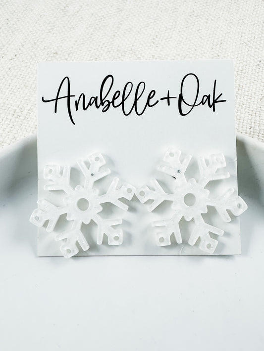 White acrylic snowflake stud earrings with subtle glitter finish, 1" total length, festive winter statement studs from Anabelle+Oak — lightweight and perfect for holiday wear.