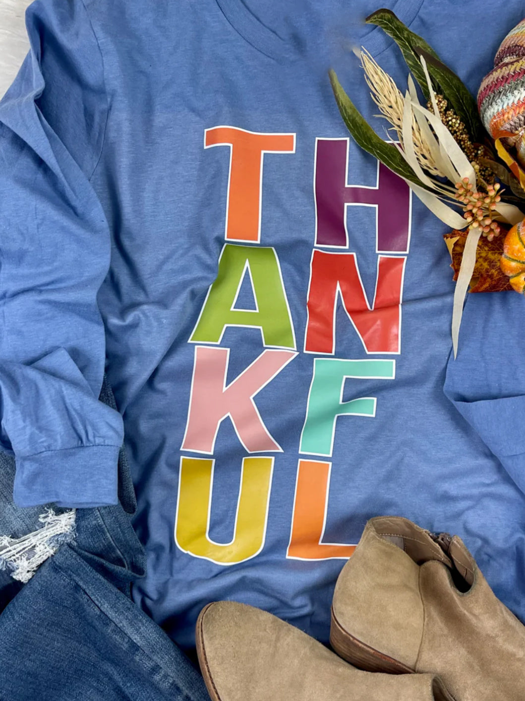 Flat lay of a blue long-sleeve tee with colorful block letters spelling “THANKFUL,” styled with tan ankle boots, distressed jeans, and a small fall floral bundle for a cozy autumn look.