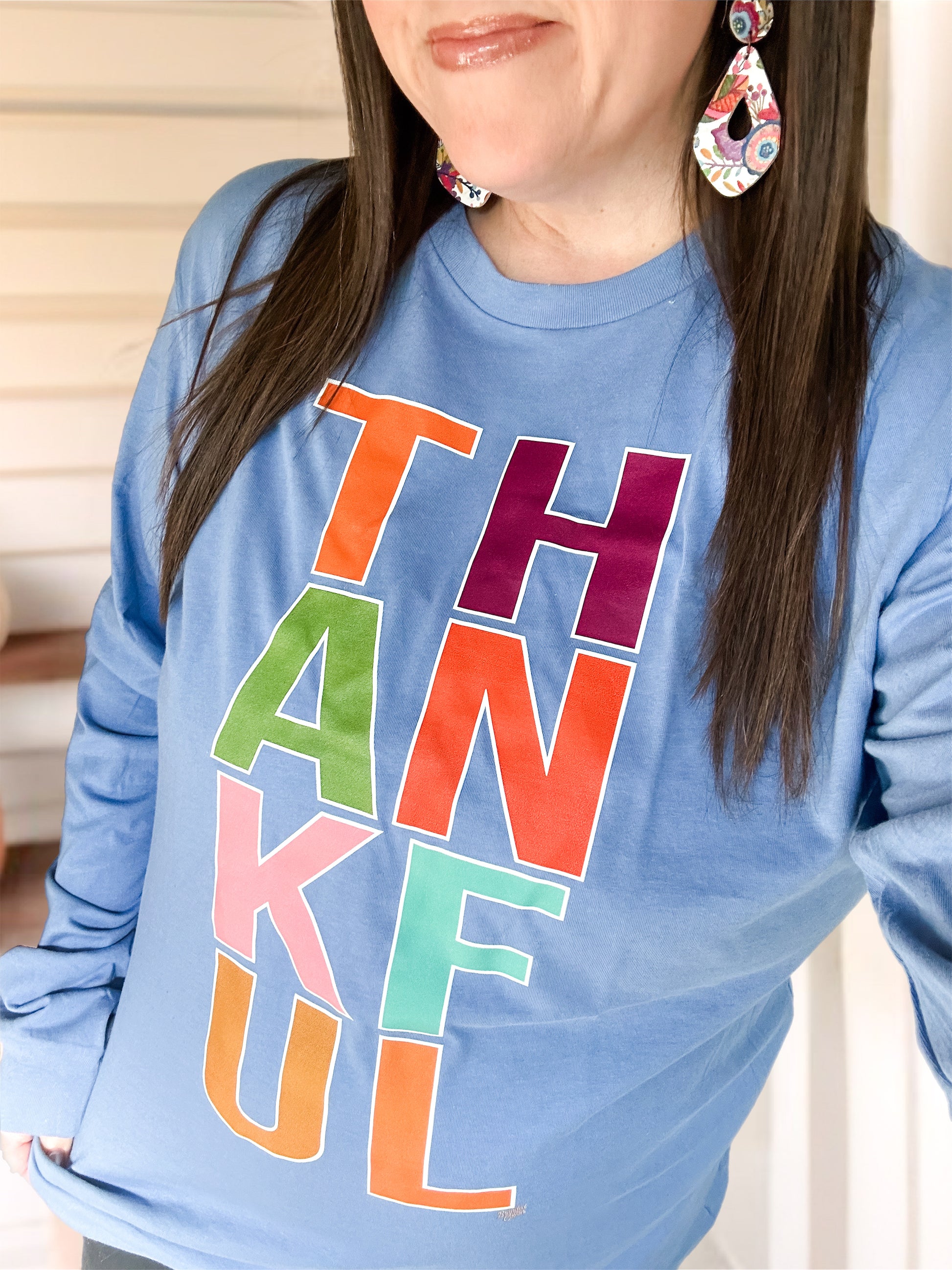 Woman wearing a blue long-sleeve tee with colorful block letters spelling “THANKFUL,” paired with bright floral teardrop earrings. The image highlights the cheerful fall design and cozy fit of the shirt.