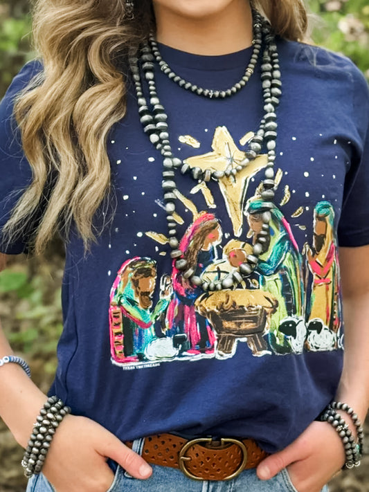 Close-up of woman wearing The Bethlehem Story Tee — navy Bella Canvas t-shirt featuring a colorful hand-painted-style nativity scene with Mary, Joseph, baby Jesus, and the Star of Bethlehem; styled with layered beaded necklaces, belt, and denim jeans.