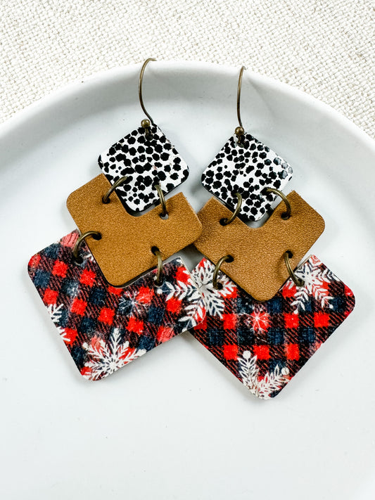 A pair of earrings featuring a mix of snowflake patterns and animal prints on a buff-colored background, displayed on a white plate.