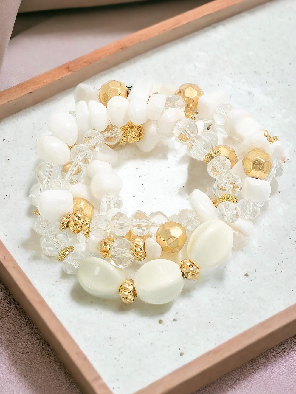 Set of white, gold, and crystal beaded stretch bracelets arranged in a circle on a white textured tray with a soft neutral background.