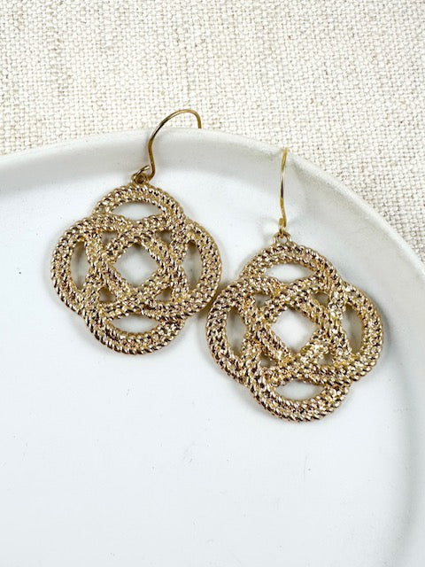 Gold woven knot earrings with a clover-shaped open design on a white ceramic plate