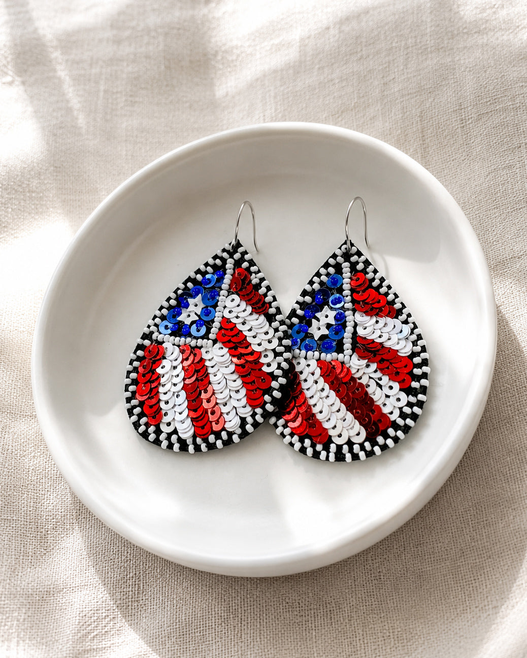 Close-up of Flag of Freedom red, white, and blue American flag earrings showcasing intricate sequin and bead details in a white ceramic dish.