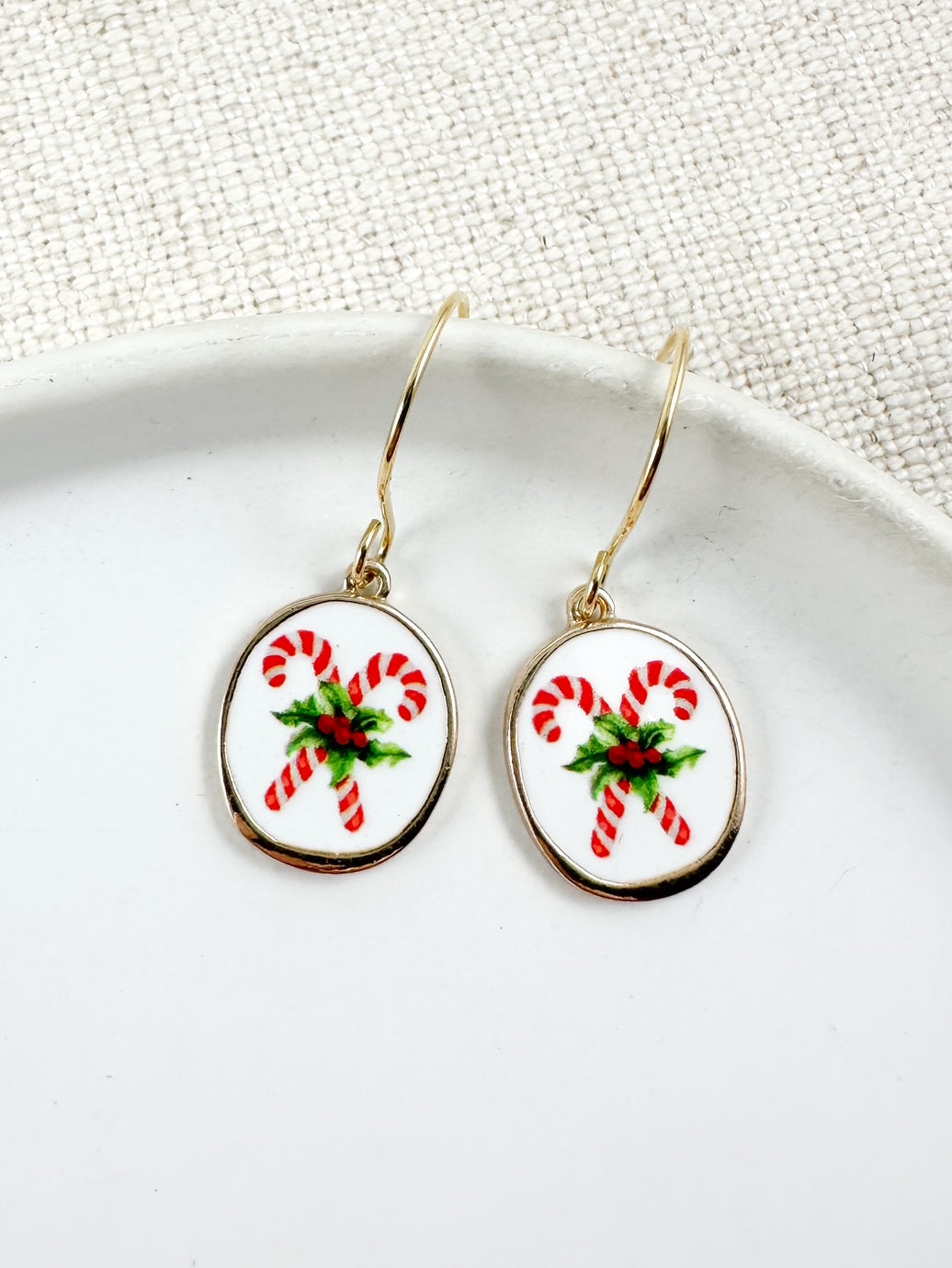Oval drop earrings with white background and red candy cane design accented with green details hanging from gold hooks on a white dish