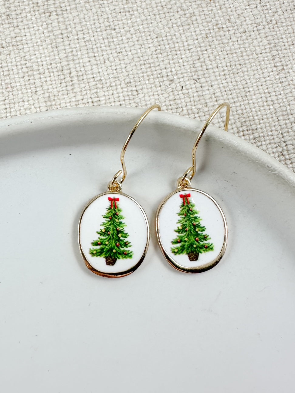 Oval drop earrings with white background and green Christmas tree design accented with small red details hanging from gold hooks on a white dish
