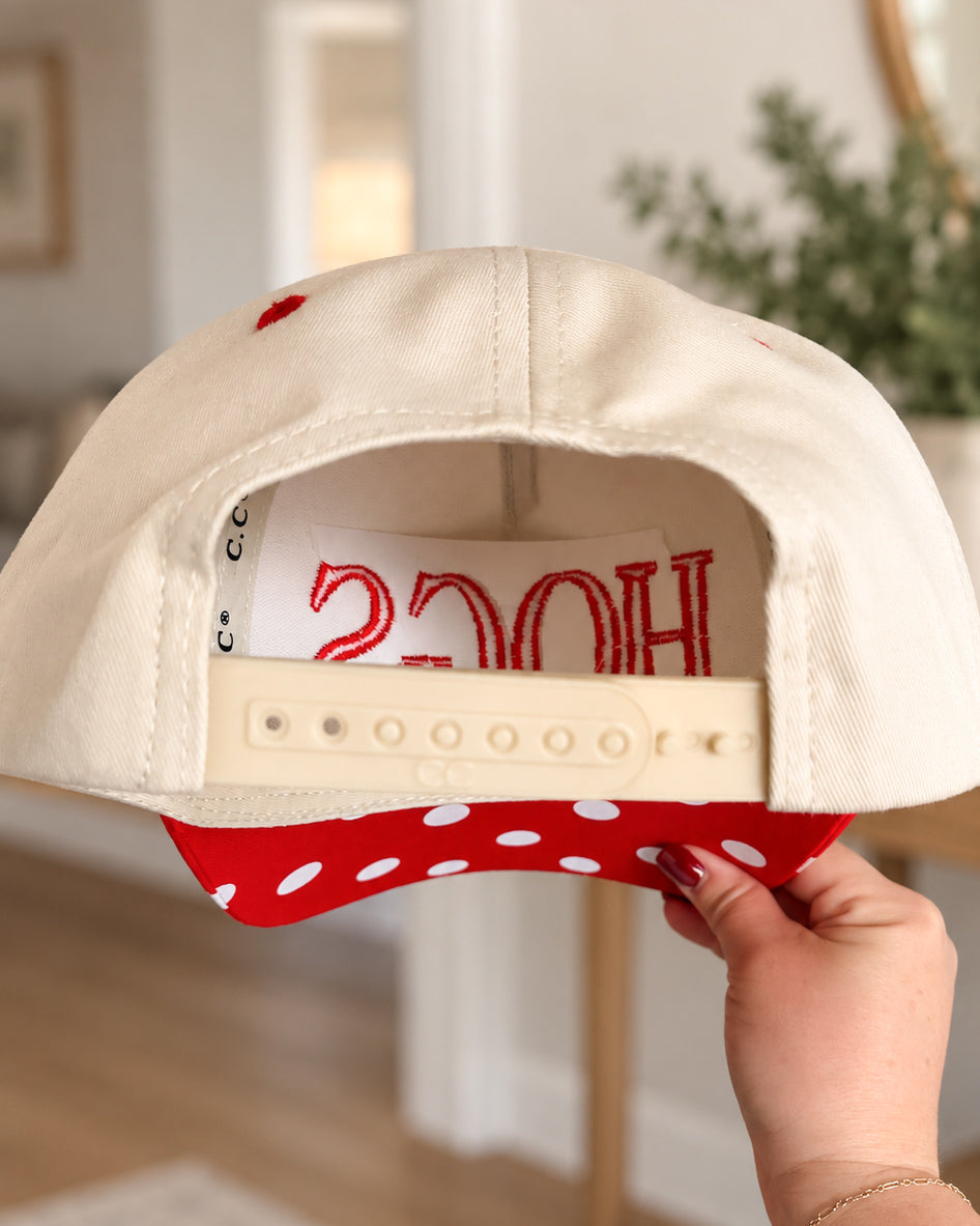 Back of the cream and red HOGS hat held in one hand, showing the adjustable snapback closure, interior embroidery, and underside of the red polka dot bill.