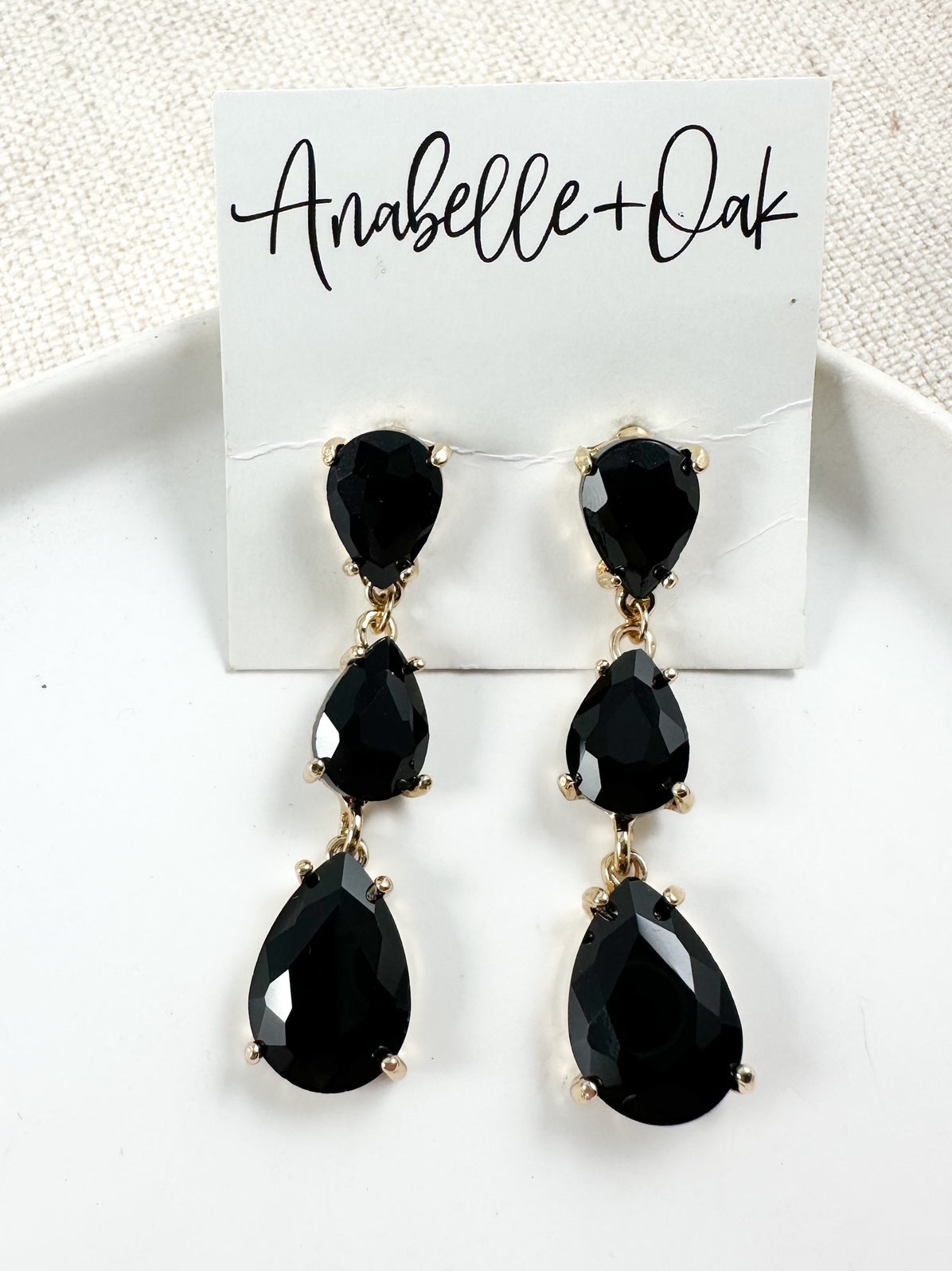 Tiered black teardrop crystal earrings with three faceted stones set in gold-tone settings displayed on a white dish