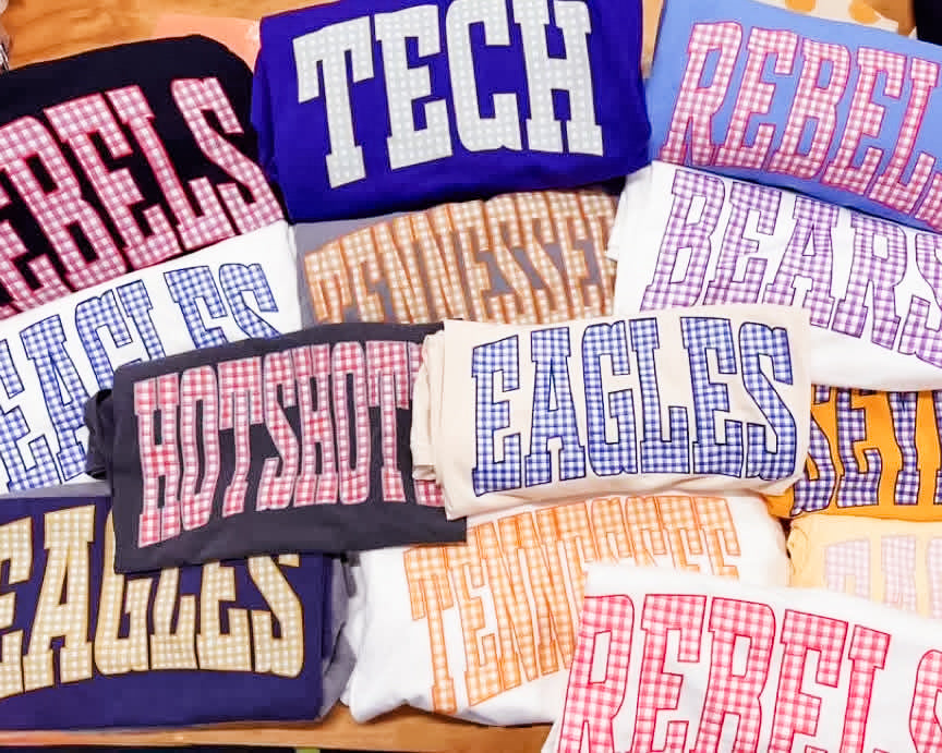 Assorted graphic tees and sweatshirts featuring oversized gingham lettering with school names and mascots in various color combinations.