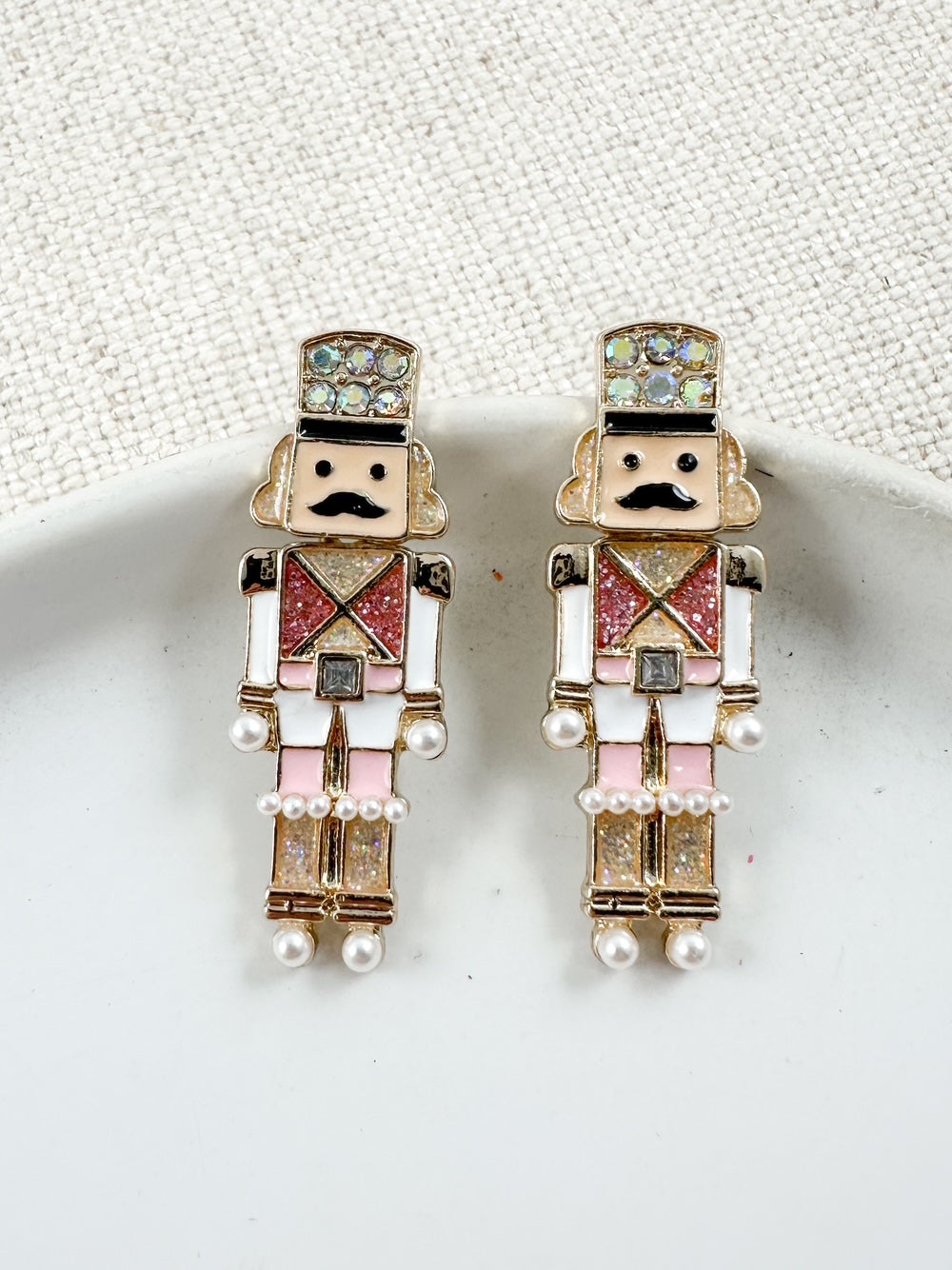 Enamel earrings shaped like Nutcracker soldiers with pink, white, gold, glitter, and pearl details, displayed on a white dish.