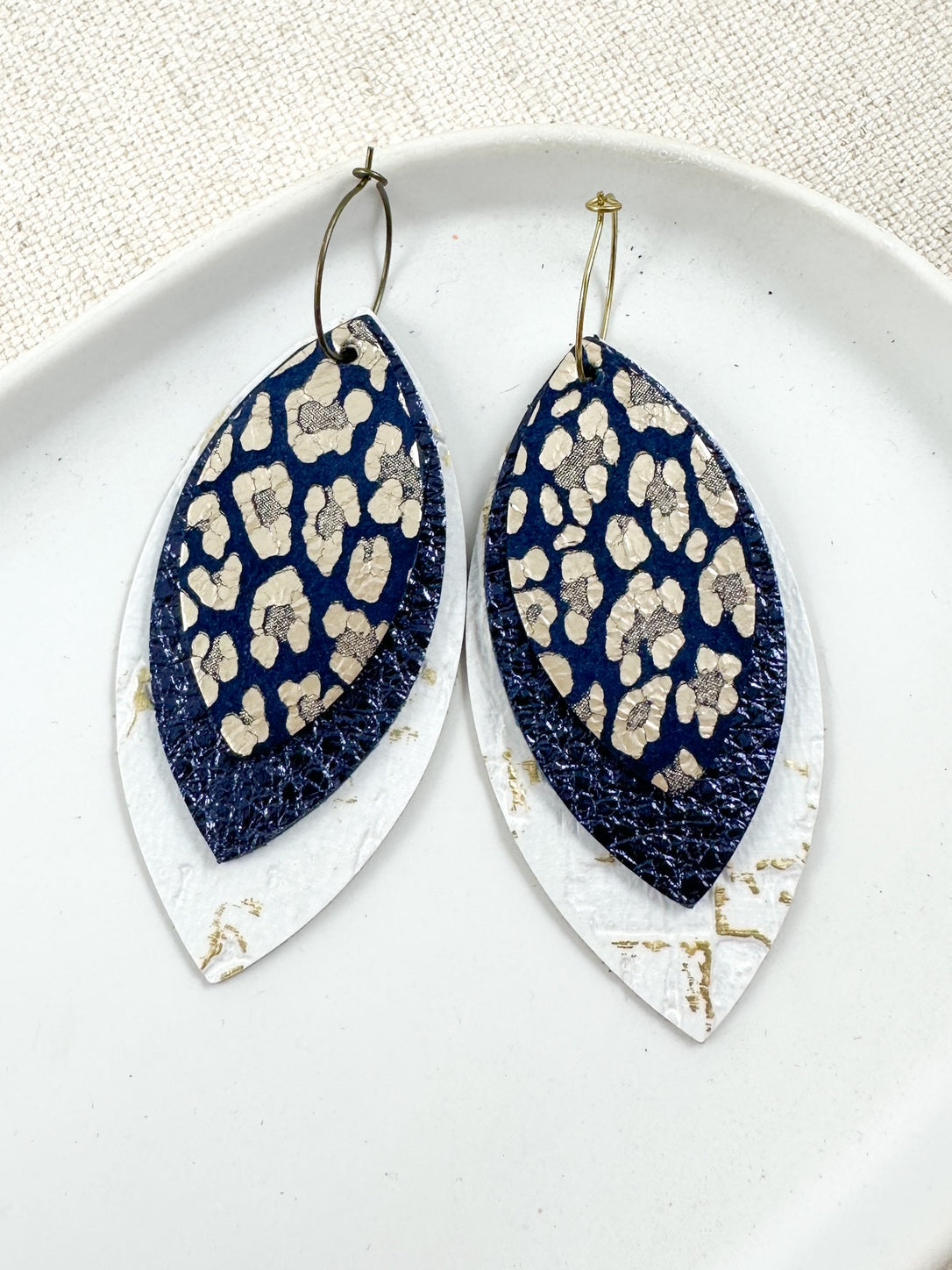 Layered navy and gold leather earrings with white distressed base, metallic navy middle layer, and navy suede top featuring soft gold leopard print, displayed on a white ceramic dish with a neutral fabric background.