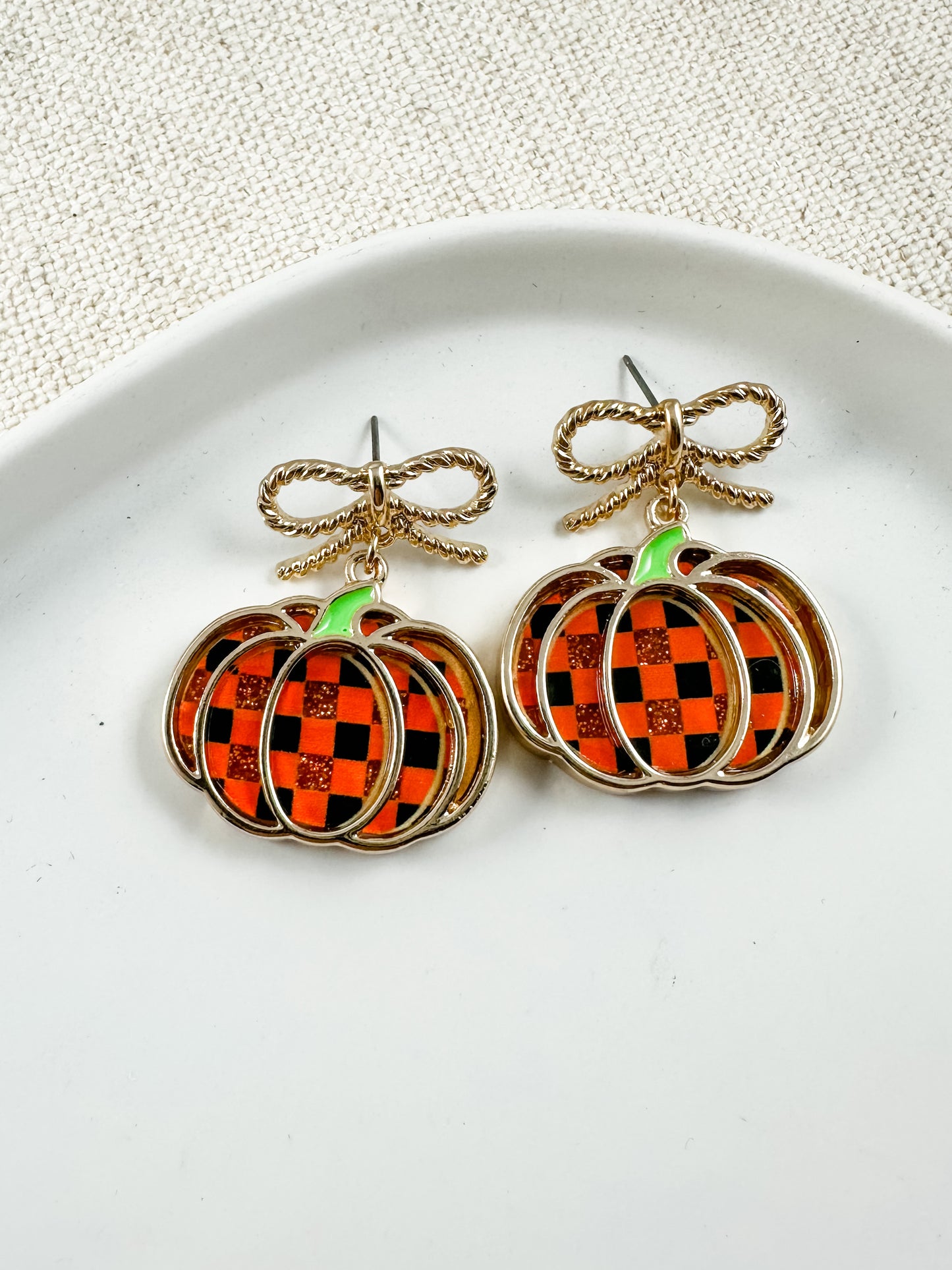 Gold bow and checkered pumpkin earrings with orange-and-black enamel details displayed on a white ceramic dish with a neutral fabric background.