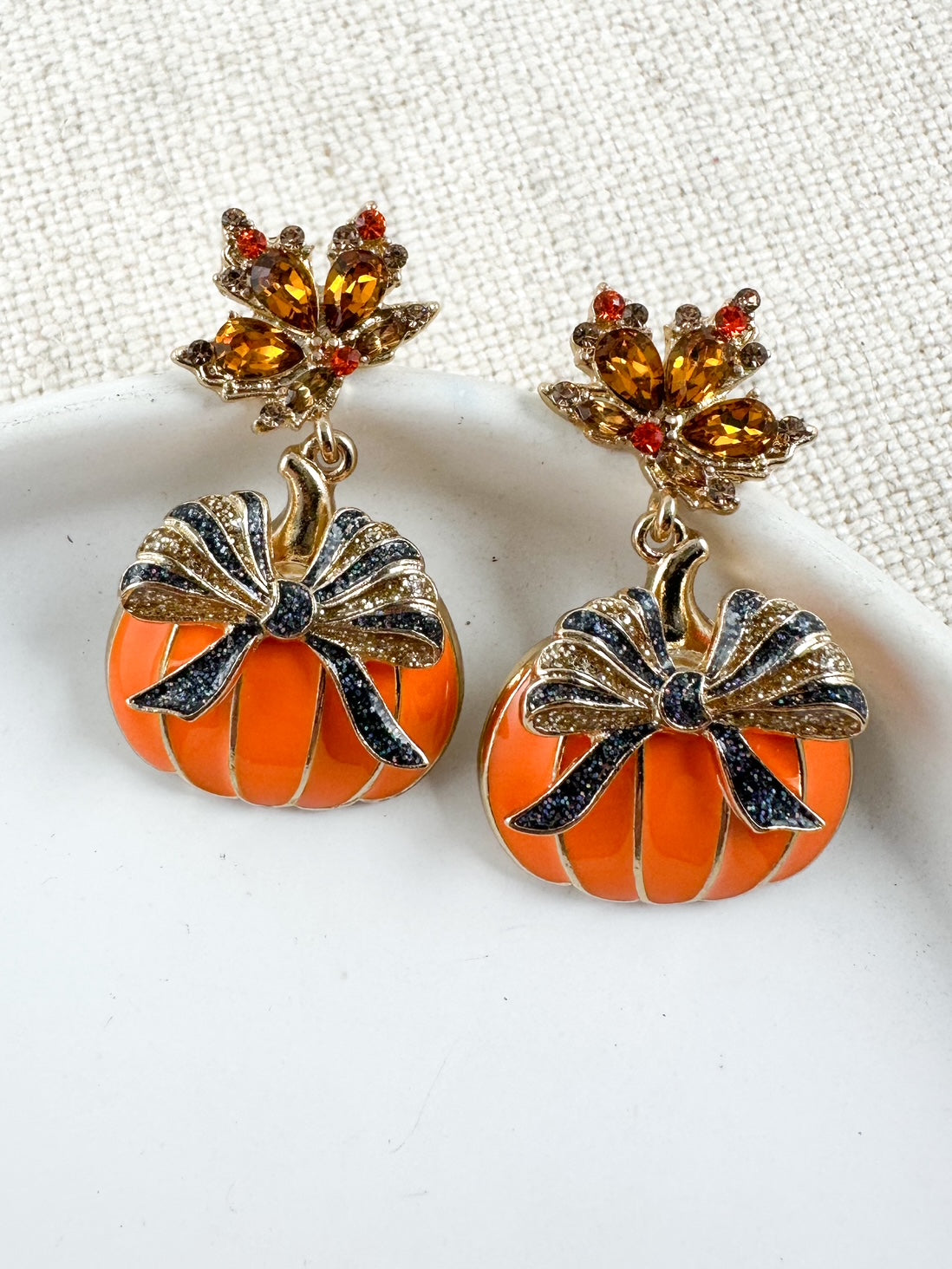 Gold pumpkin earrings with brown glitter bows and amber gemstone leaf tops displayed on a white ceramic dish with a neutral fabric background.