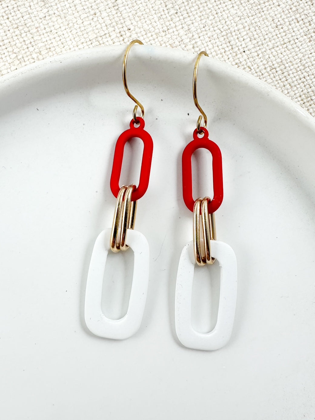 Linked drop earrings with red and white geometric links and gold accent rings displayed on a white ceramic dish with a neutral fabric background.