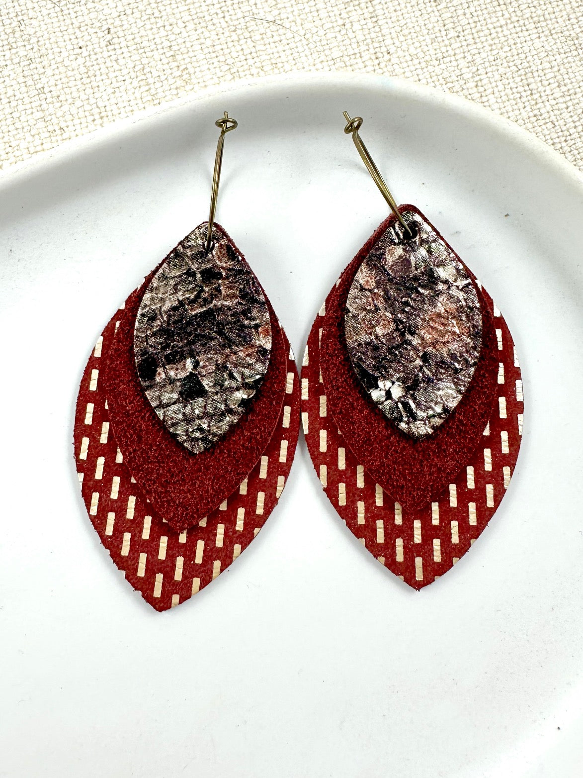Layered maroon, gold, and snake print leather earrings with suede texture, displayed on a white plate.