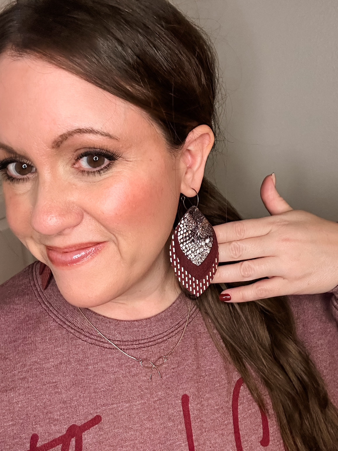 Woman wearing maroon layered leather earrings with gold and snake print details, styled with matching maroon nails and a heather maroon “Thankful” sweatshirt.