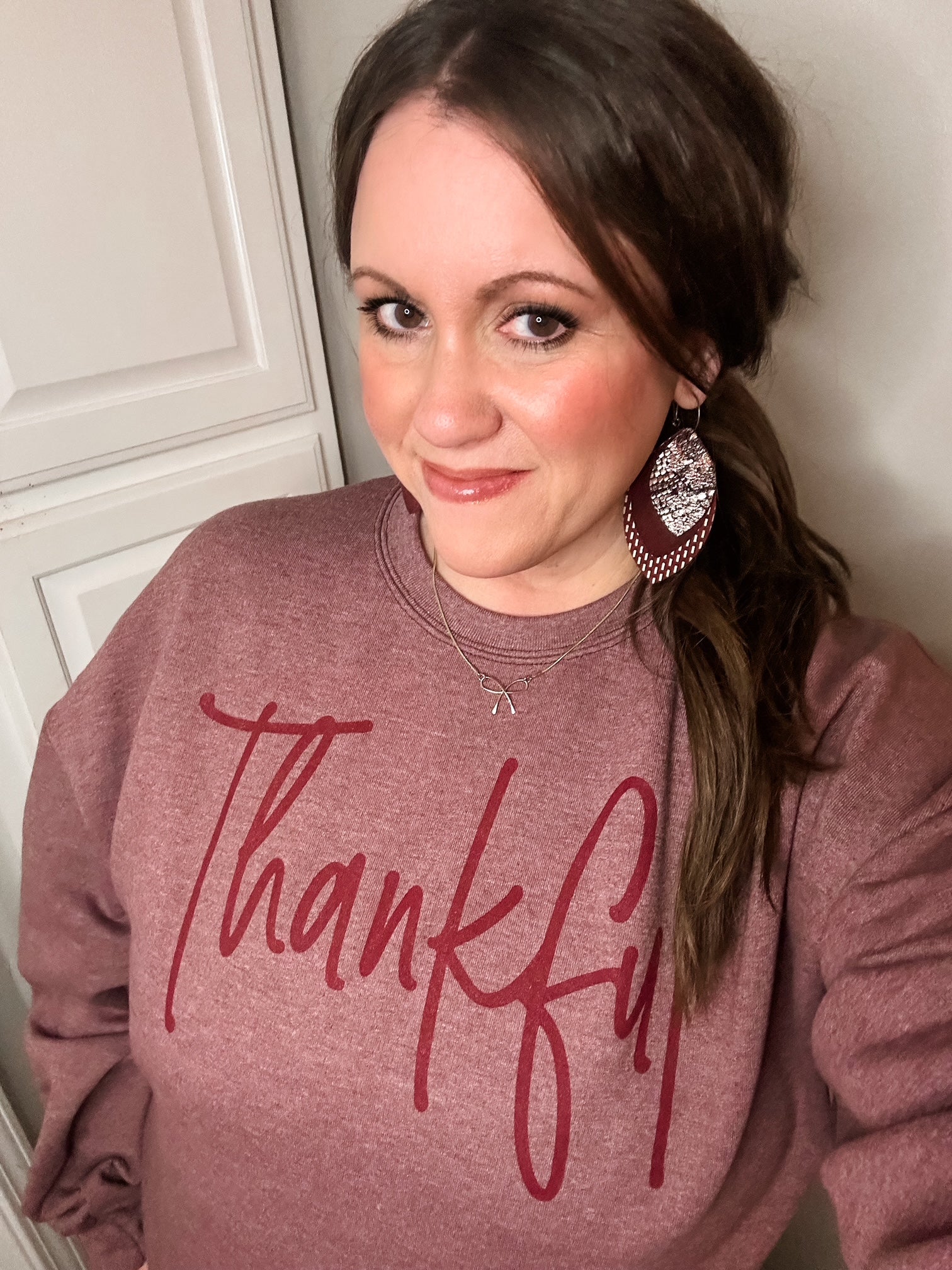 Woman smiling and wearing a heather maroon “Thankful” sweatshirt with red script lettering, paired with large patterned earrings and soft, natural makeup.