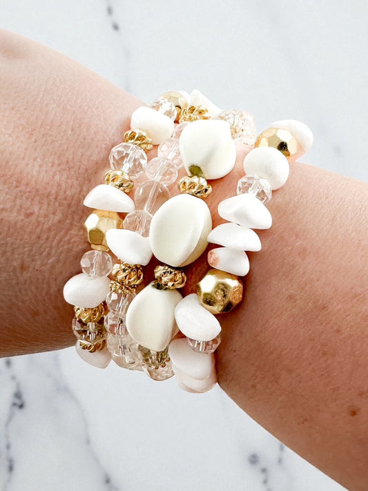Stack of four white, gold, and crystal beaded stretch bracelets worn on wrist against a marble background.
