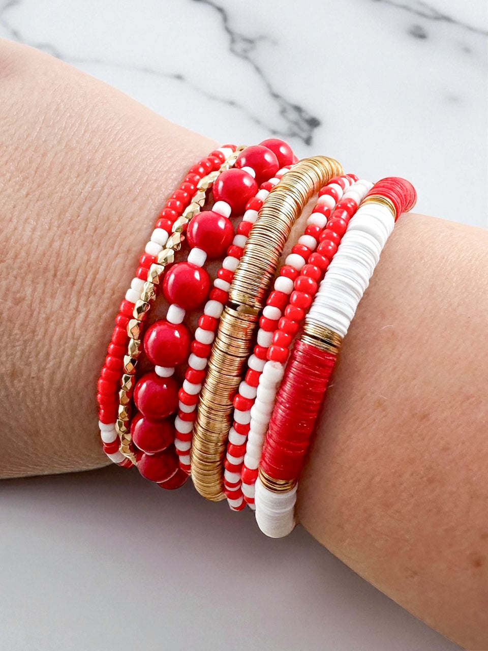 Set of eight red, white, and gold beaded stretch bracelets featuring heishi, seed, and metal beads worn stacked on a wrist against a marble background.