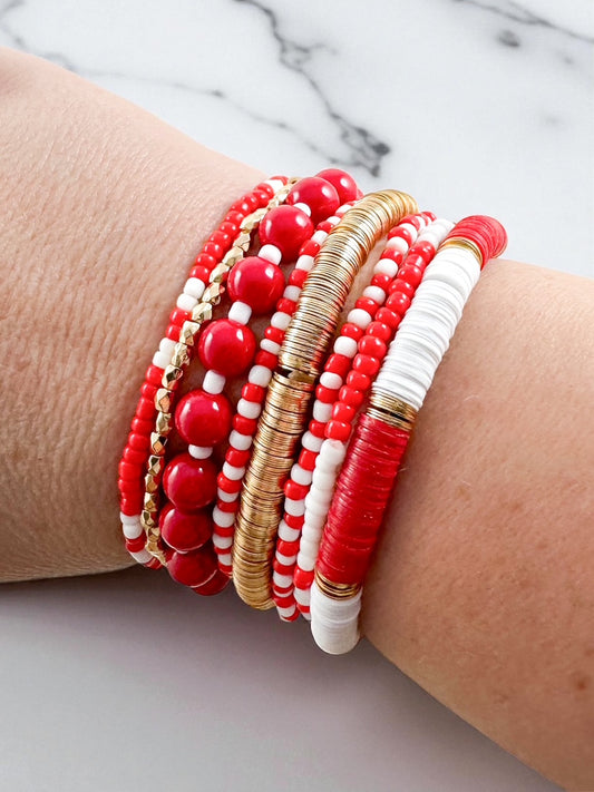 Set of eight red, white, and gold beaded stretch bracelets featuring heishi, seed, and metal beads worn stacked on a wrist against a marble background.