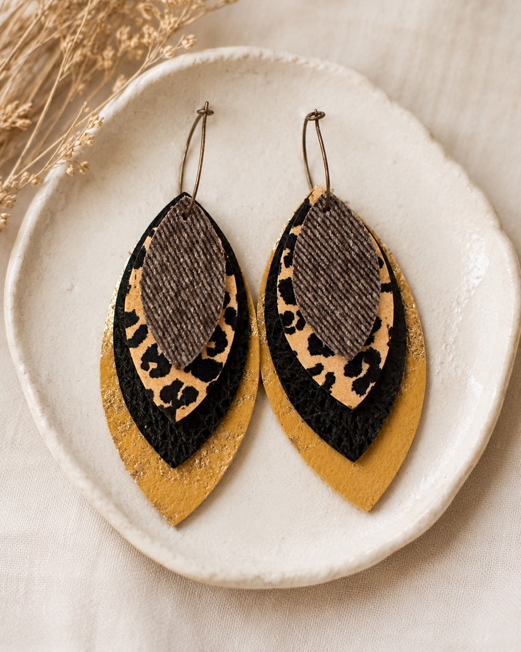 Layered leopard, black, textured brown and metallic gold earrings displayed together in a simple white jewelry dish.