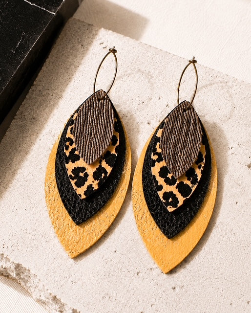 Layered leopard, black, metallic gold and textured brown leather earrings styled on a light stone surface with black accents.