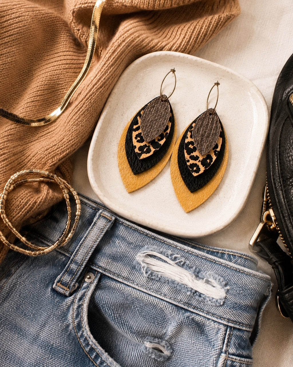 Leopard, black and metallic gold statement earrings styled with medium-wash denim, camel and black textures in an outfit flat lay.