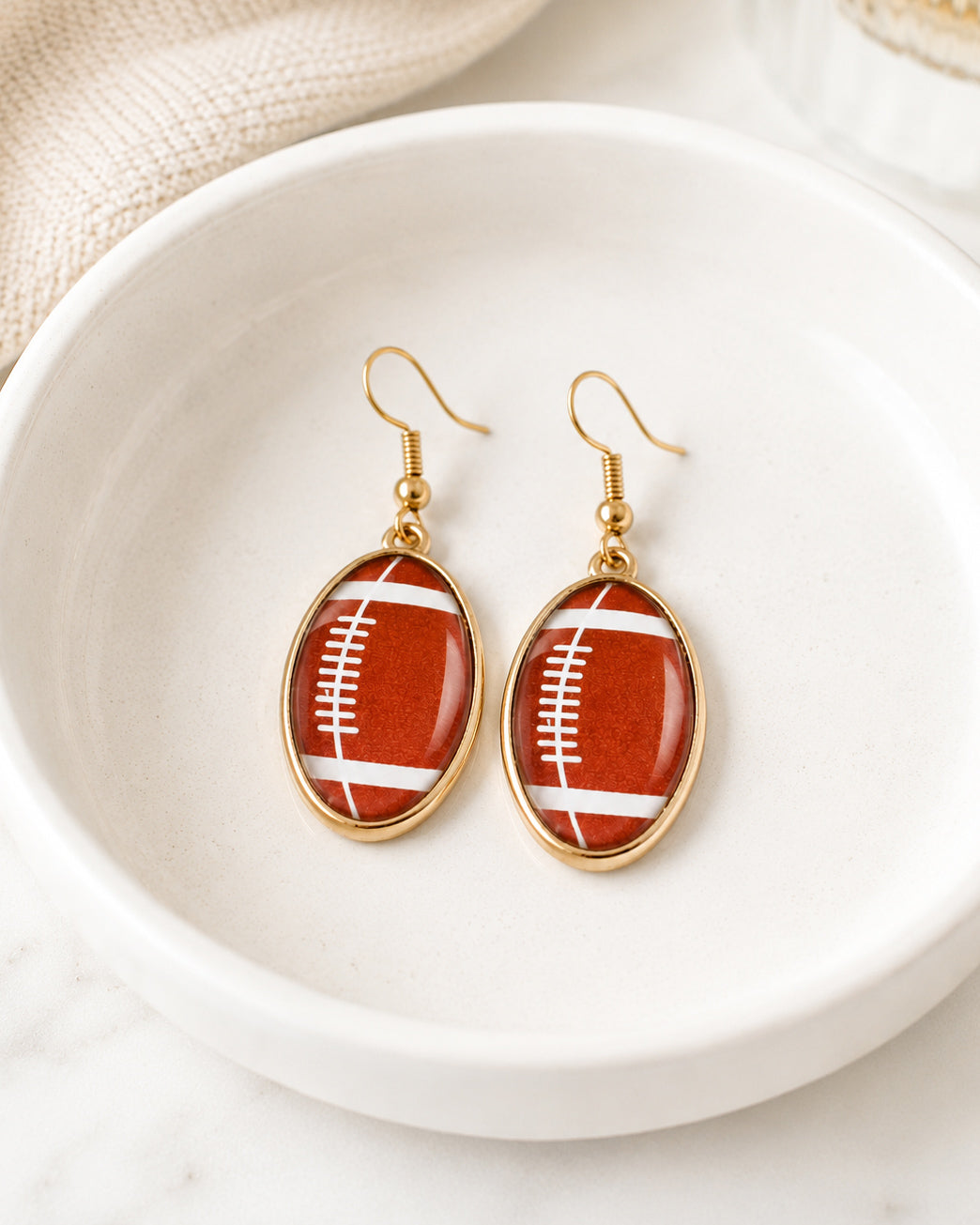 Gold football dangle earrings displayed in a white ceramic jewelry dish.