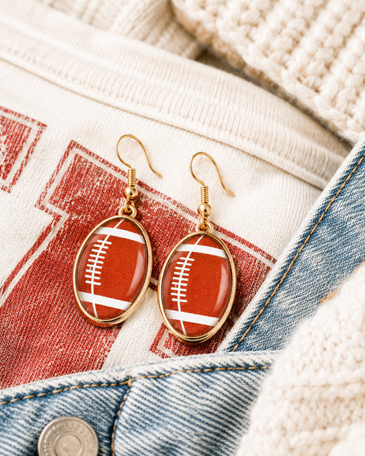 Gold football dangle earrings styled with a cream football graphic tee, denim jacket, and cozy knit sweater.