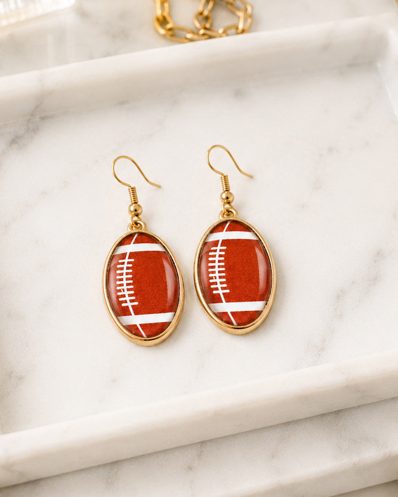 Gold football earrings displayed on a white marble tray in natural light.