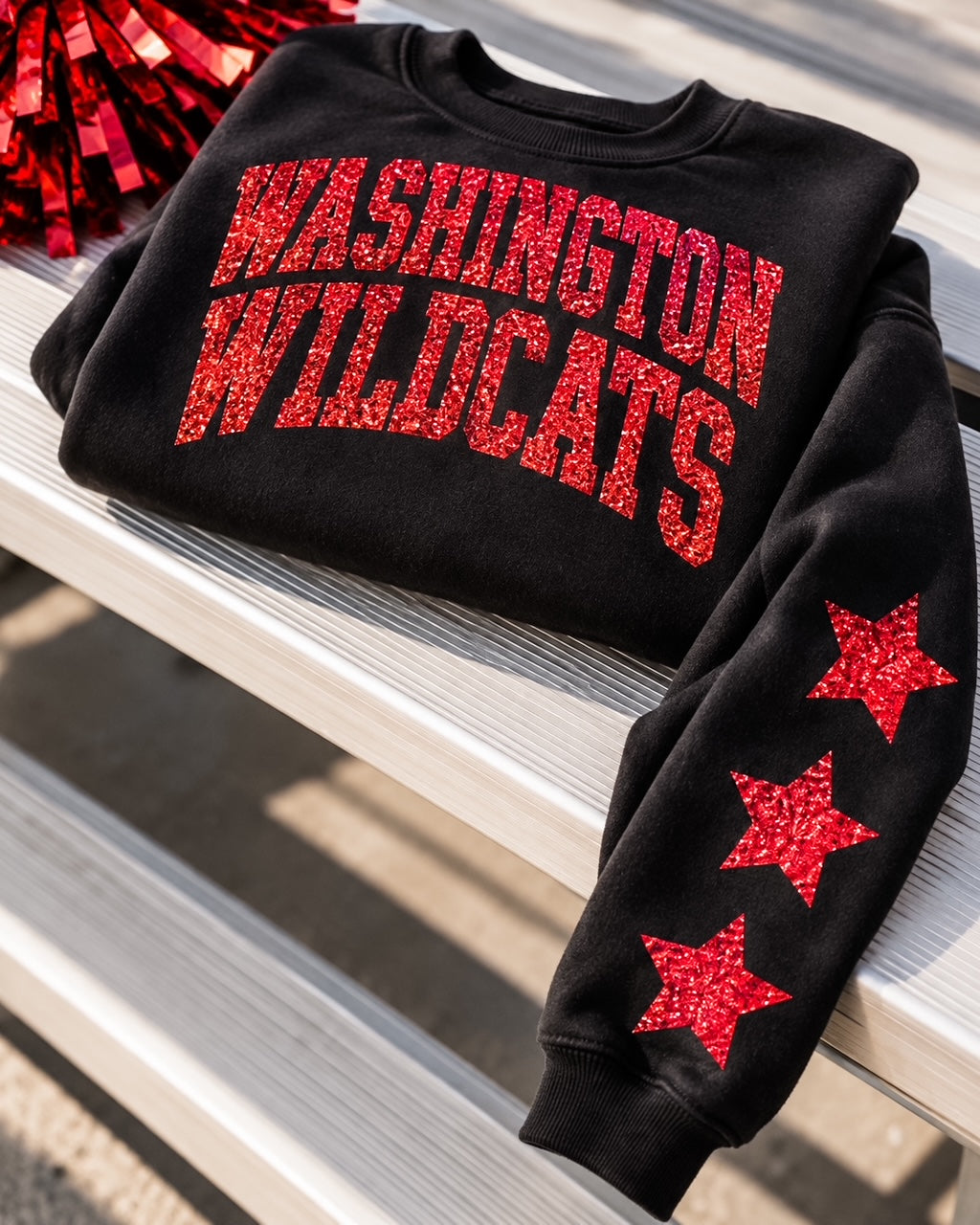 Black Washington Wildcats glitter sweatshirt photographed in a school bleacher setting, featuring red glitter lettering and sleeve stars.