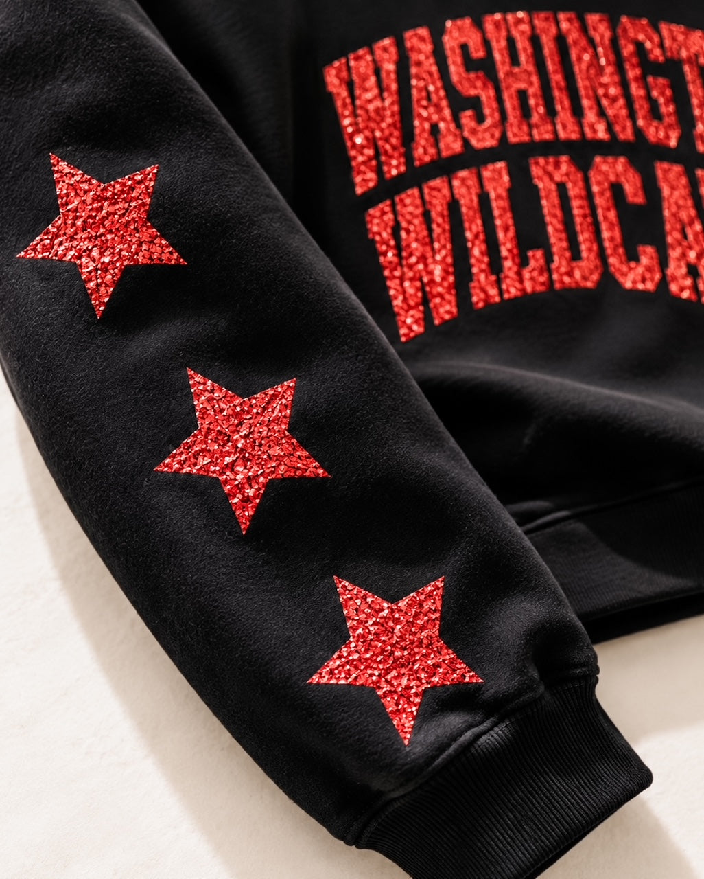 Close-up of red glitter Washington Wildcats lettering and coordinating glitter sleeve stars on a black crewneck sweatshirt.