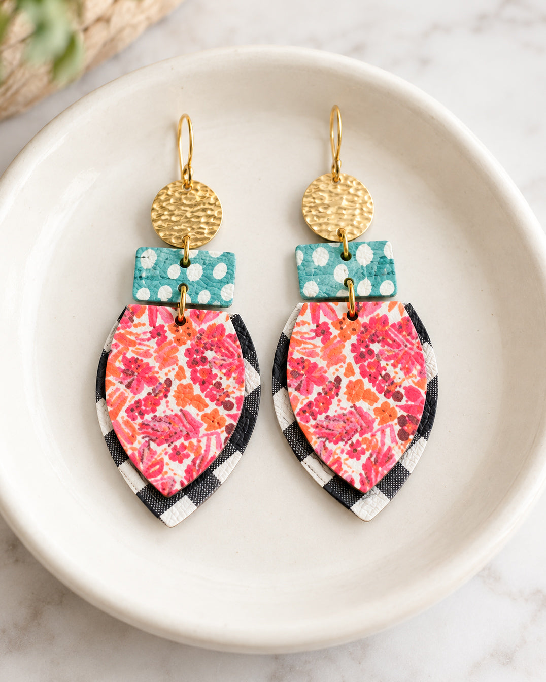 Pair of All the Pretty Things statement earrings displayed in a shallow ivory ceramic dish, featuring hammered gold circles, turquoise polka dot rectangles, and layered pink floral and black-and-white gingham leather drops.