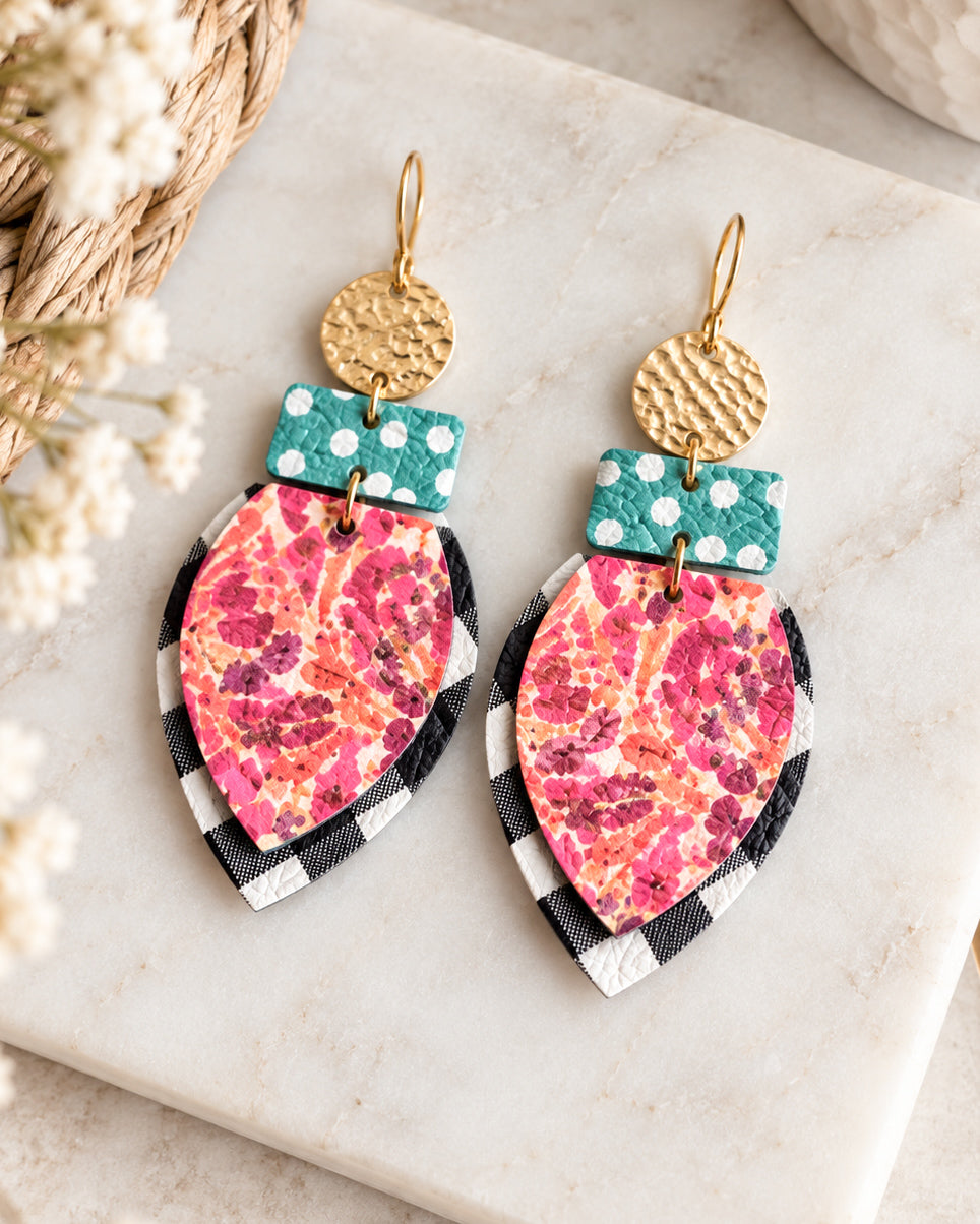 All the Pretty Things handmade earrings arranged on a cream marble surface beside a woven basket and dried flowers, featuring pink and coral floral leather, turquoise polka dots, black-and-white gingham, and hammered gold circles.