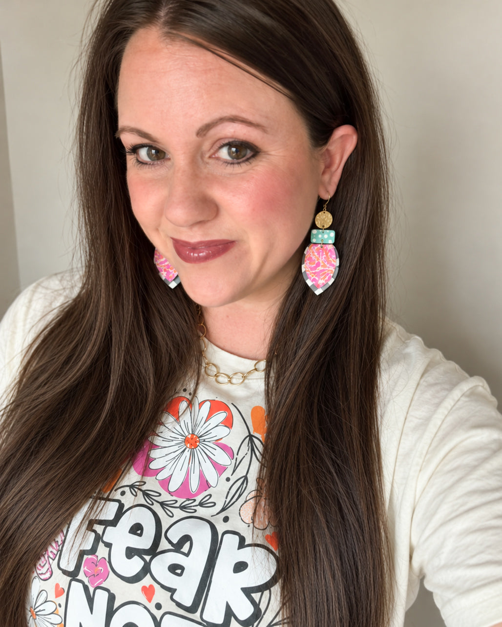 Woman with long brown hair modeling All the Pretty Things colorful statement earrings with pink floral, turquoise polka dot, and black-and-white gingham layers, styled with a cream floral graphic tee and gold chain necklace.