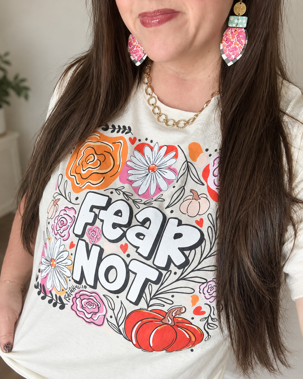 Woman wearing colorful handmade leather and cork statement earrings with pink and orange florals, turquoise polka dots, black-and-white gingham, and hammered gold accents, styled with a cream Fear Not floral graphic tee and gold chain necklace.