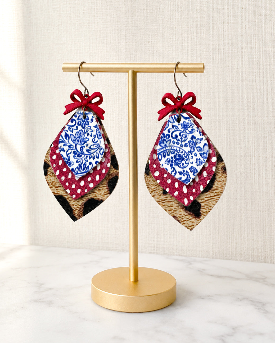 American Honey Earrings displayed on a matte gold T-stand, featuring layered genuine leather and cork with red bows, blue and white floral print, red polka dots, and leopard print accents.