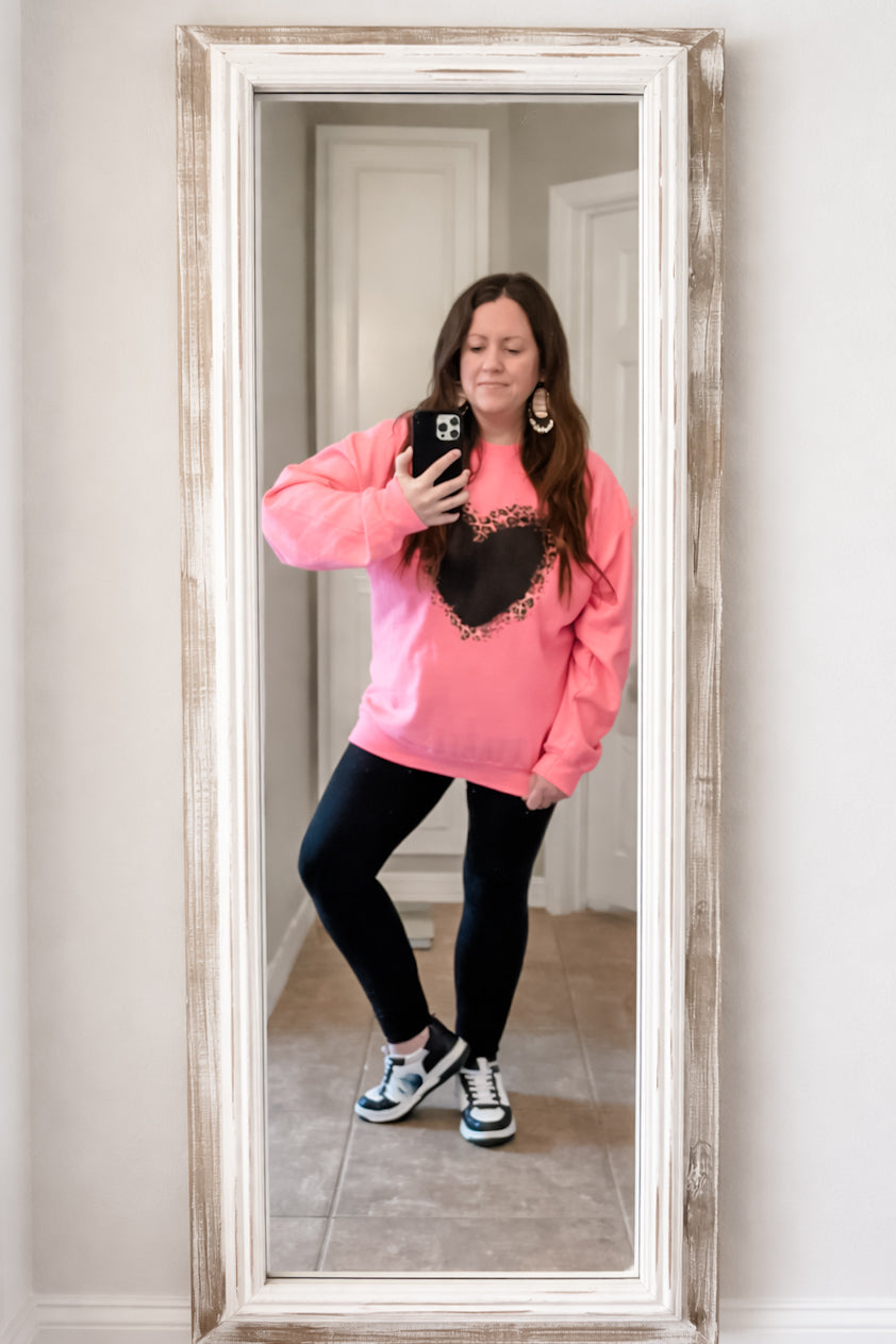 Woman wearing a bright pink sweatshirt with a blackwashed leopard heart graphic, styled with black leggings and sneakers, taking a mirror selfie indoors