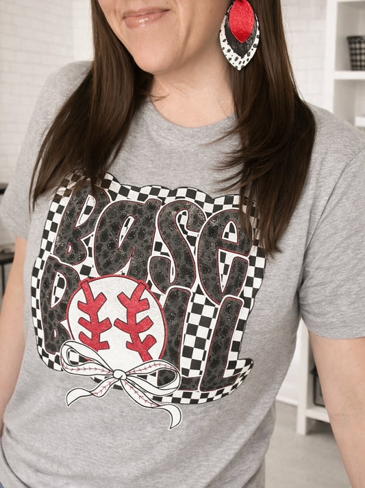 Woman wearing heather gray baseball graphic tee with checkerboard leopard lettering and red baseball bow design with red layered earrings.