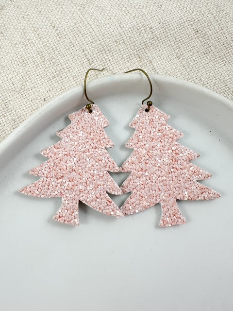 Close up of pink glitter Christmas tree shaped earrings with matte sparkle, hanging on antique bronze hooks on a white dish.