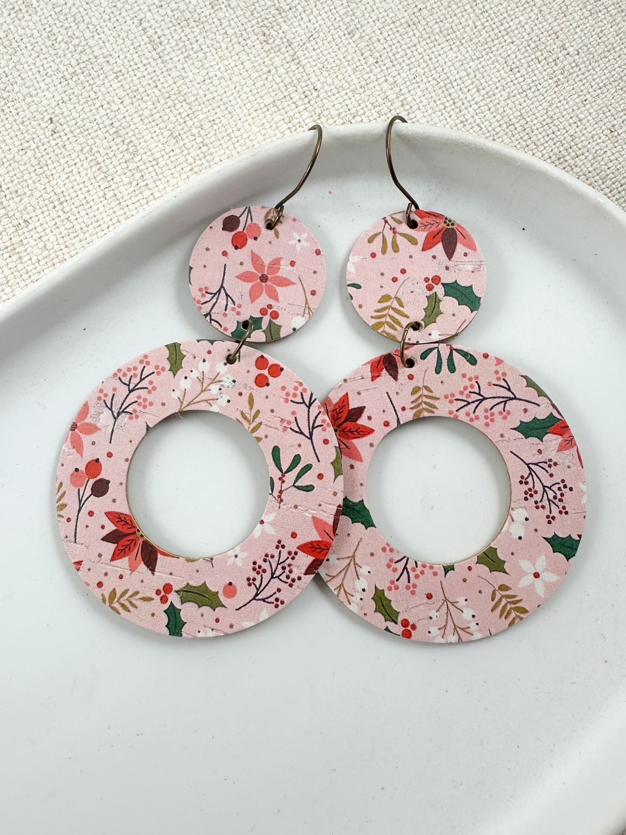 Blush Poinsettia Cork circular earrings displayed on a white plate with a linen-textured background, featuring the pink poinsettia and holiday berry pattern.