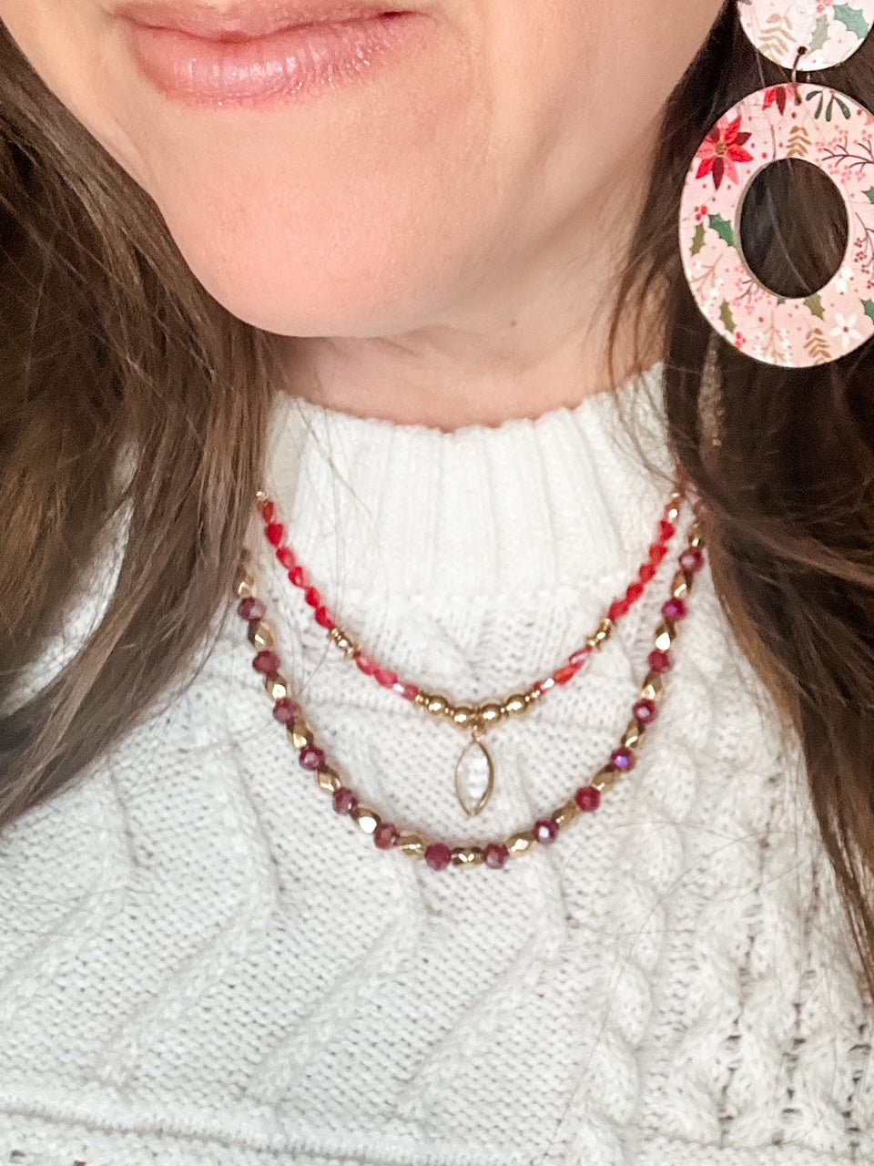 Model wearing Blush Poinsettia Cork circular earrings with a white cable knit sweater and layered red and gold beaded necklaces.