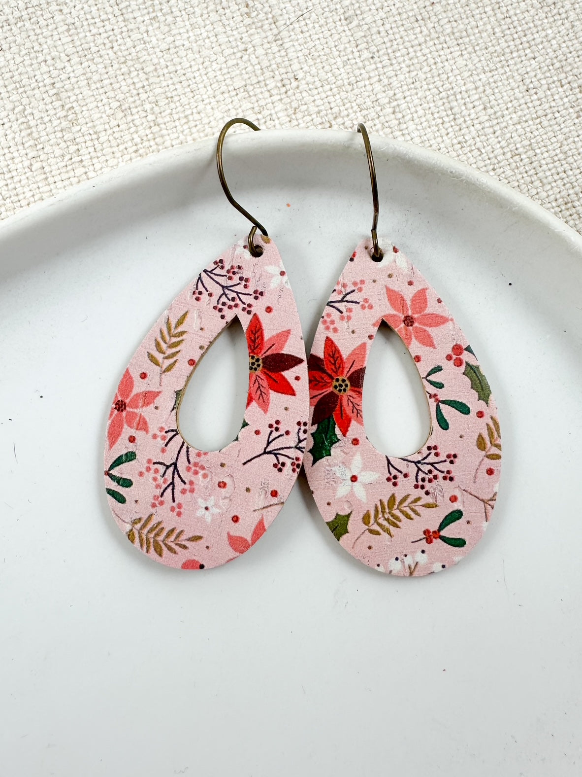 Blush Poinsettia Cork teardrop cutout earrings displayed on a white plate with a linen-textured background, featuring pink poinsettias, berries, and greenery.