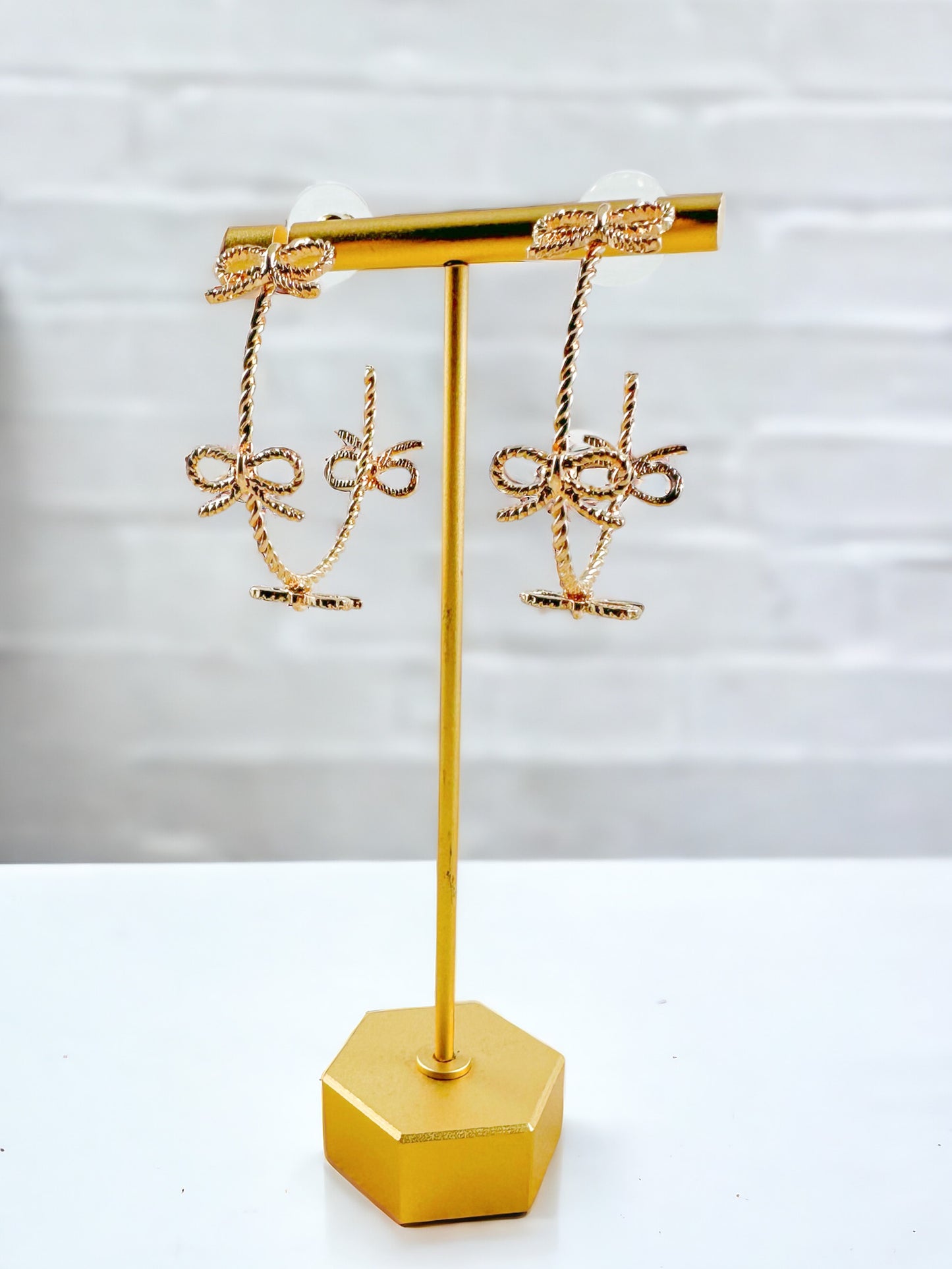Gold hoop earrings with small twisted metal bows around the hoop displayed on a gold stand