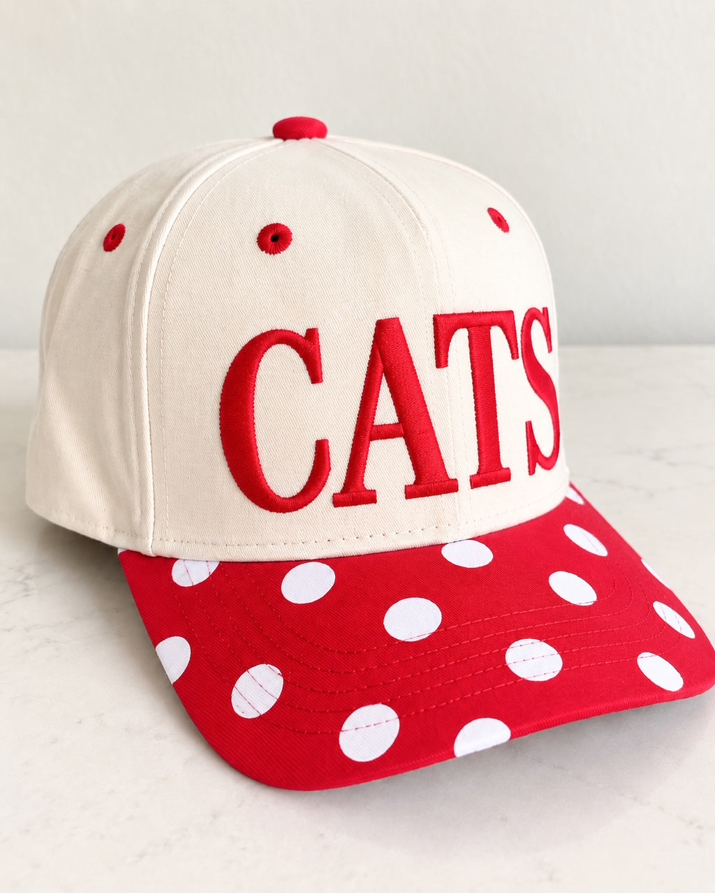 Angled view of cream CATS Pep Rally Hat showing red embroidery, red eyelets, and the polka-dot bill.